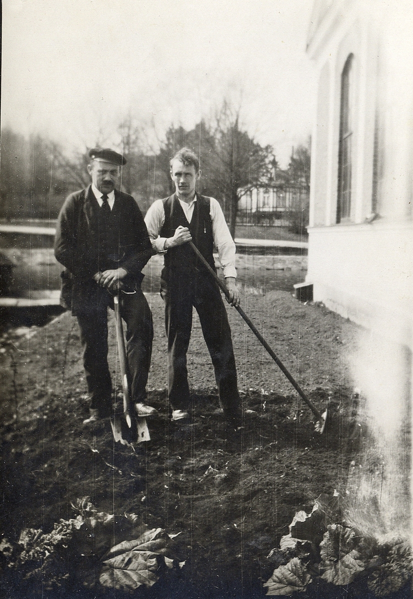 Två män, bröderna Johan och Edvin Åström, håller på med trädgårdsarbete utanför elverket i Växjö. I bakgrunden skymtar Växjösjön och dåv. järnvägsrestaurangen.