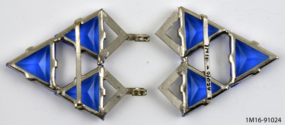 Skärpspänne av metall med glaspärlor, två delar. Sex stycken triangelformade blå glaspärlor fästade mot metall. Prismärkning: N.a.g. kr. 12:50.