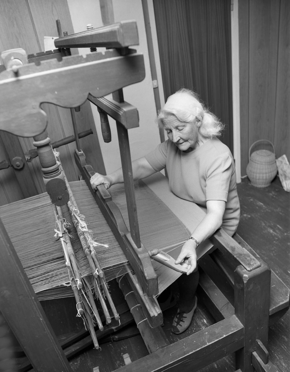 Gunnhild Vestad (1906-1994) fra Trysil, fotografert ved vevstolen i andreetasje i hjemmet sitt, der hun satt og produserte stoff som hun seinere skulle sy klær av. Gunnhild giftet seg i 1929 med Lemmek Vestad (1901-1977) og sammen bygde de bruket Vangen i Slettås-grenda, hvor de ryddet 130 dekar jord og drev mjølkeproduksjon. Lemmek hadde inntekter fra skogsarbeid i vintersesongen. Da var det Gunnhild som tok ansvar for de seks ungene, buskapen og mangt annet. Kontantsinntektene var beskjedne, så det gjaldt å være sjølforsynte med det meste. Arbeidet ved vevstolen inngikk i strevet for å begrense pengebruken, og mange av tekstilene Gunhild vevde ble etter hvert til arbeidsklær for ektemannen.