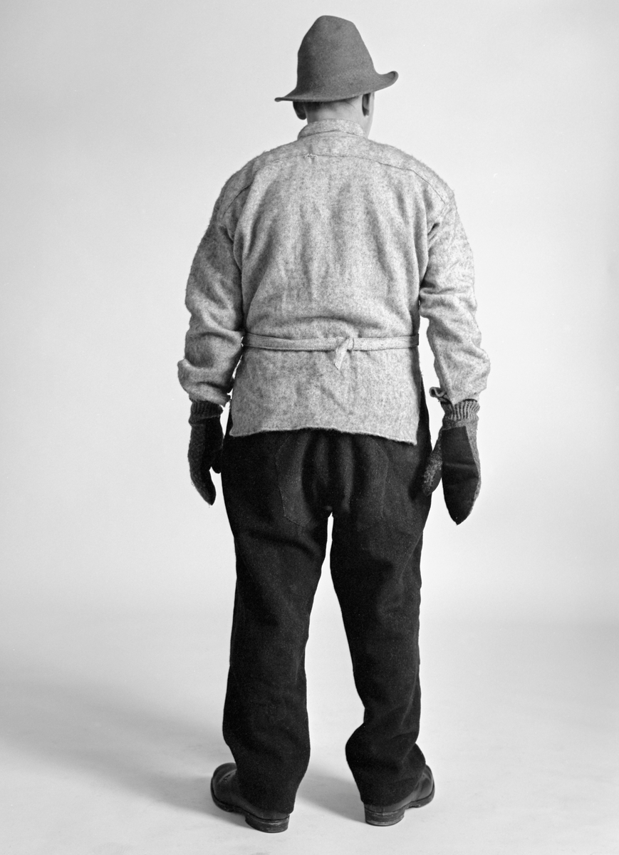 Demonstrasjon av hvordan en skogsarbeider i mellomkrigstida kunne være kledd når han var klart til å gå ut i vinterskogen og starte arbeidsdagen. Bildet er tatt bakfra, altså mot ryggsida. Dette er et studioopptak, gjort på Norsk Skogbruksmuseum med autentiske klær, og med skogsarbeiderveteranen Konrad Eggen (1917-1992) fra Elverum som modell. Bildene ble tatt med tanke på Geir Hovensjøs artikkel «Skogsarbeiderklær», som skulle publiseres i museets årbok. 

Modellen hadde en filthatt på hodet, sannsynligvis enm utrangert finhatt. Fordelen med hatten var at den hadde en brem som gav beskyttelse mot regn og mot snødryss når skogsarbeideren begynte å hogge i rotsona, slik at barmassen kom i bevegelse. Hatten var mye brukt under hogst og fløting, men ettersom den ikke dekte ørene var det nok enkelte tømmerkjørere som foretrakk andre hodeplagg, i hvert fall når vendingene ble lange. På overkroppen hadde modellen en busserull av halvull, atankelig med ei skjorte under. På spesielt kalde dager gikk det an å ha en ullgenser - en «islender» - mellom skjorta og busserullen. Men hogsten var tungt fysisk arbeid, der kroppen utviklet mye varme. Derfor forekom det nok også, i moderate vintertemperaturer, at tømmerhoggeren vrengte av seg det ytre laget og arbeidet bare i skjorta. På hendene har modellen strikkevotter med innlagt vadmelsstoff i grepet. Harde grep om øksesklaft og saghandtak slet nemlig hardt på vottene. På underkroppen hadde modellen ei romslig, mørk vadmelsbukse. Dette stoffet hadde den fordelen at fukt fra regn og snø hadde vanskelig for å trenge gjennom. Slik unngikk skogsarbeideren å fryse. Modellen på bildet hadde lærstøvler på beina, sannsynligvis beksømstøvler.
