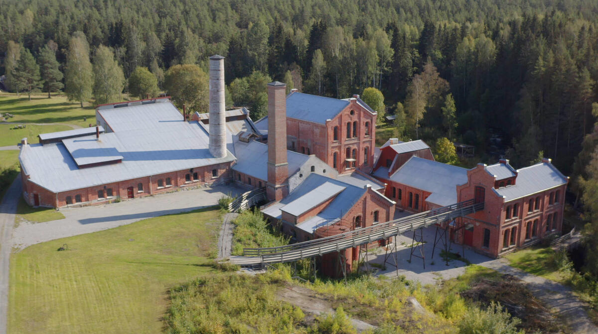 Klevfos industrimuseum sett fra lufta. Stor mursteinsbygning med to store fabrikkpiper. Sommerkledd landskap med grønne plener og frodig skog i bakgrunnen.