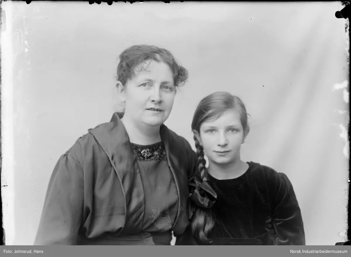 Portrett av to kvinner sittende i studio. Begge kledd i kjole, den ynste med flettet hår og knytet sløyfe i bunn.