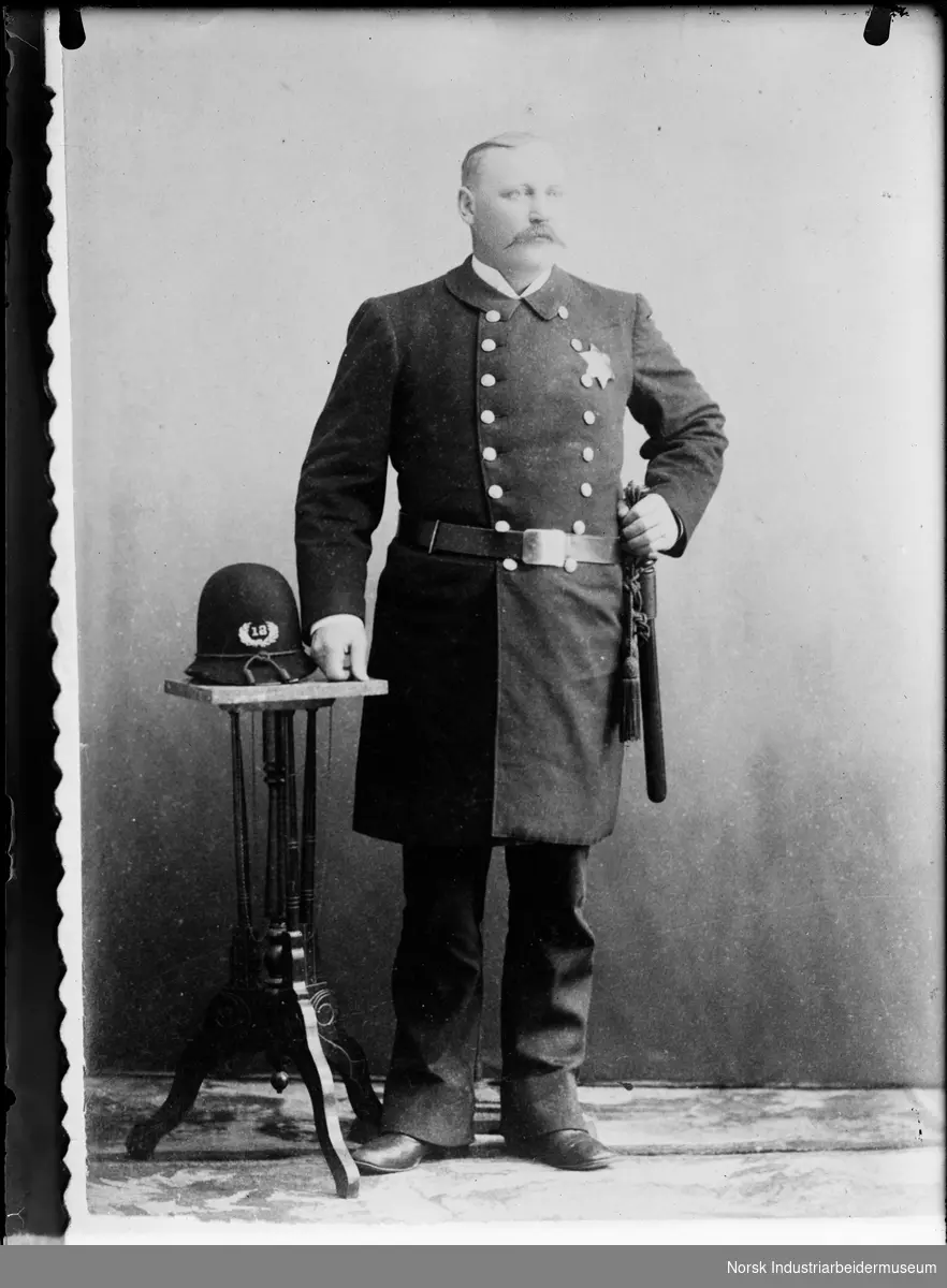 Mann i politiuniform med stjerne på venstre side av brystet. Dobbelspent knelang frakk og belte med batong på venstre side. Hjelm merket 12 med tråd rundt bremmen liggende på bord på høyre side.