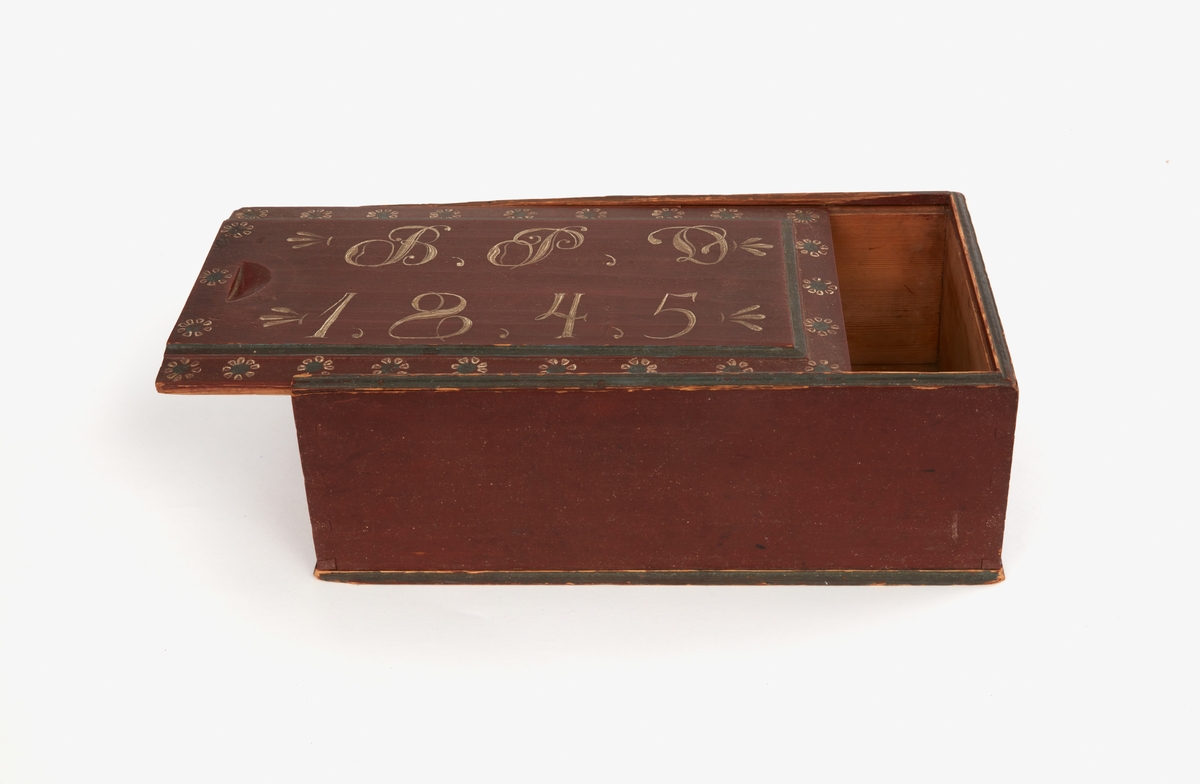 Timpask av trä. Rektangulär låda med skjutlock. Sinkade hörn, pluggad botten. Rödbrunmålad, på locket stiliserade blommor och "BPD, 1845" i grönt och vitt.