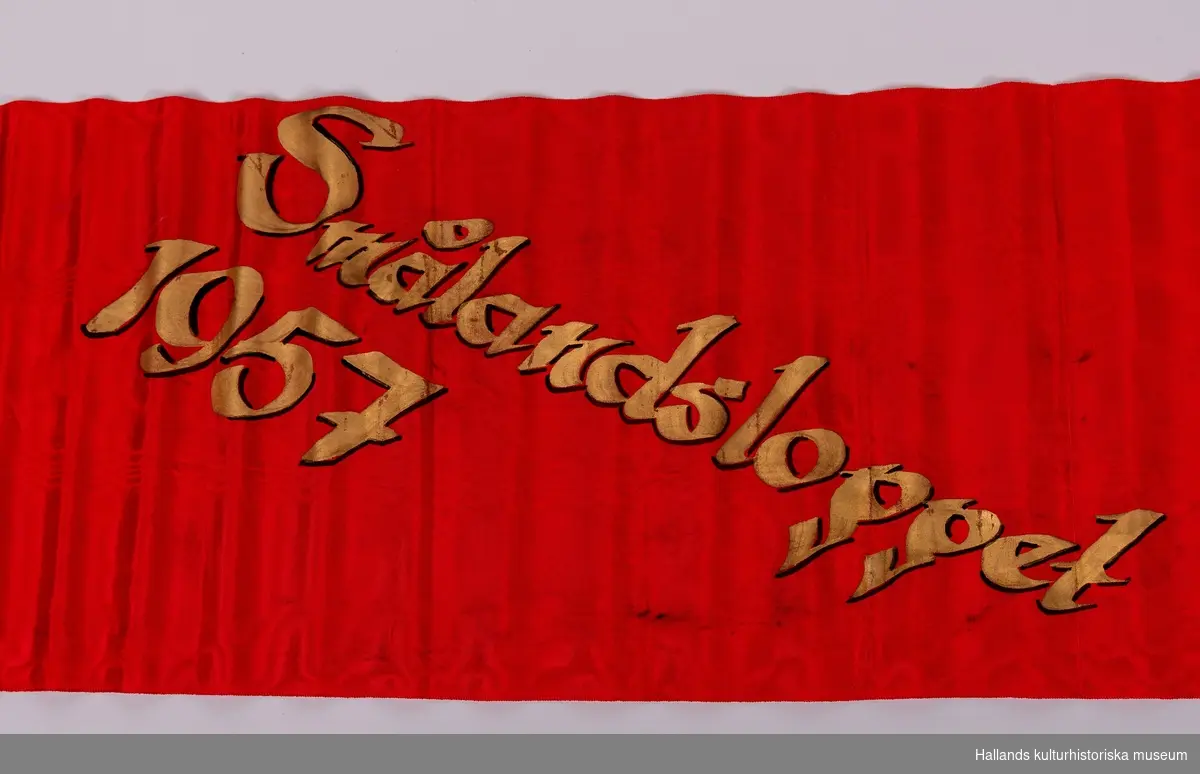 Rött konstsidenband med målad guldtext: "FONALÖBET 1957".