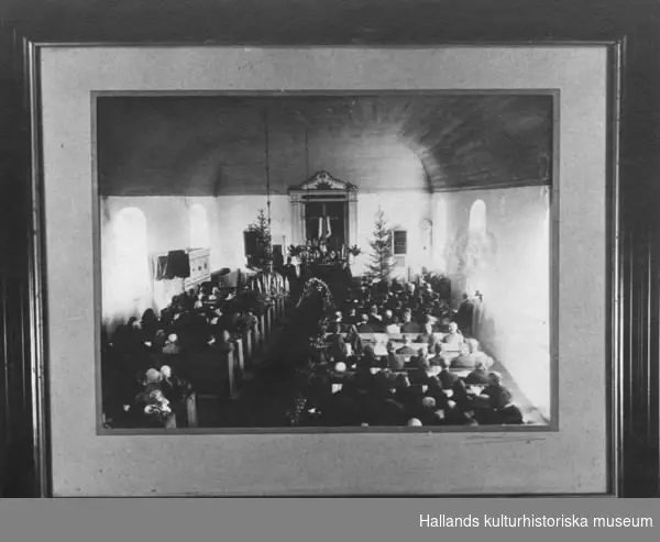 Fotografi inom ram. Föreställande begravning i Drängsereds kyrka. I koret den smyckade kistan. Bänkarna fyllda av människor. Kvinnorna huvudsakligen på vänster sida, männen till höger (syd). Kyrkan prydd med två granar samt björkris. Troligen från 1920-talet. Inramad i en brun träram, guldkant närmast bilden.