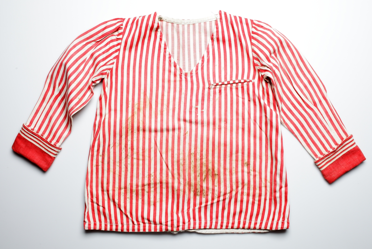 Röd- och vitrandig bomullsskjorta i sjömansmodell för barn, med långa ärmar, V-ringad halsöppning och dragsko med bomullsband längs nederkanten. Axelsömmarna är öppna med knäppning av tryckknappar i metall. Ytterligare tryckknappar inuti halsringning för att fästa sjömanskrage i. Påsydda röda manschetter med tre smala påsydda bomullsband i var ärmlinning. Insydd bröstficka till vänster (utan kantning).