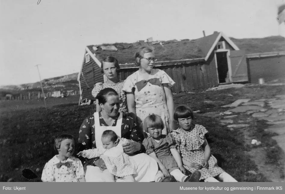 Marie Sigfrida Normann sitter omringet av sine sine barn Astrid, Ragnhild og Kari samt tre av deres søskenbarn fra Berlevåg foran en låve på Melkøya i Finnmark i sommeren 1935.  Søskenbarna er barn av formann Johansen på moloen, dette ifølge giveren.