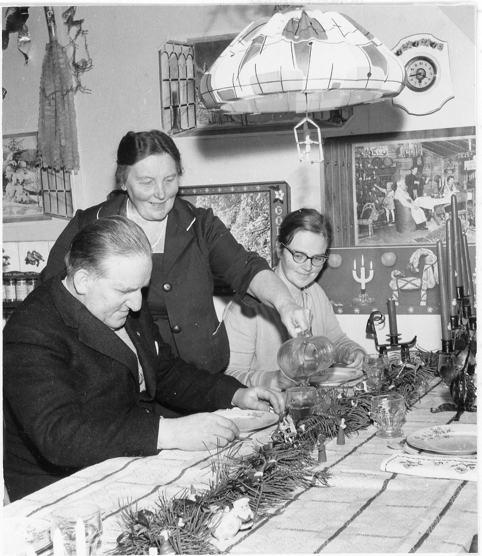 En man och en yngre kvinna sitter vid ett dukat bord, en kvinna står mellan dem och häller upp dryck från en kanna. Det är jultid, med bland annat 5-armad ljusstake på bordet och julbonad på väggen.