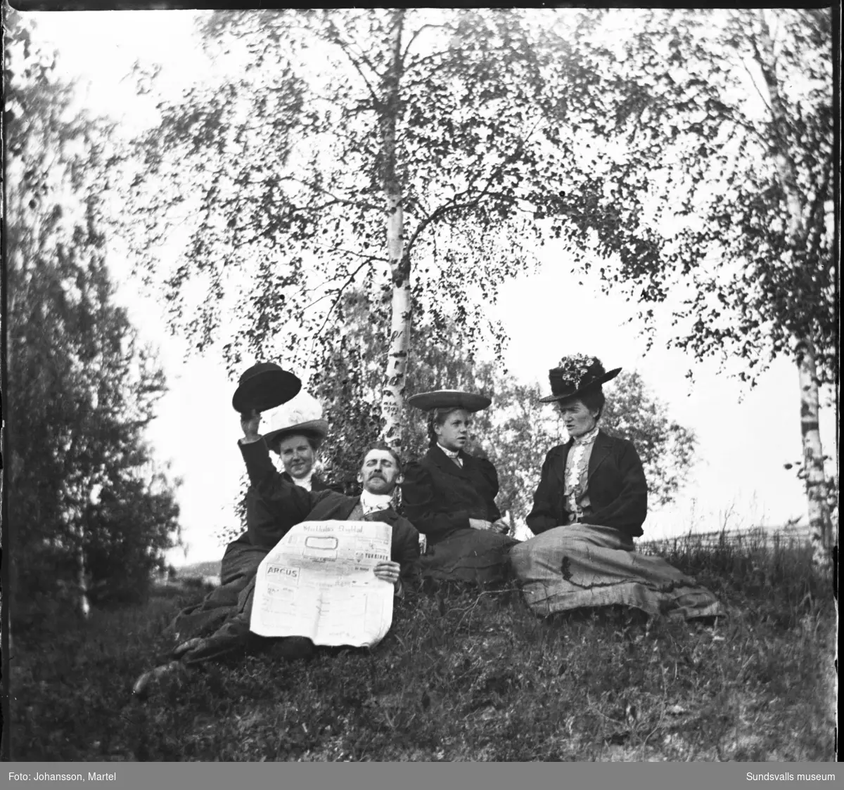 Tre kvinnor med stora hattar samt en man med en tidning, Stockholms Dagblad, sitter i det gröna.
