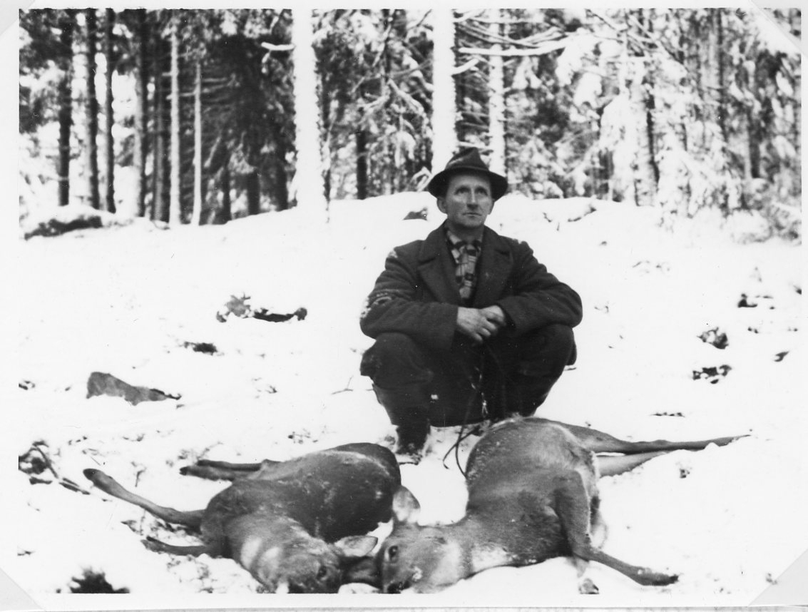 En man med hatt och rock sitter på huk i snön bakom två skjutna rådjur.