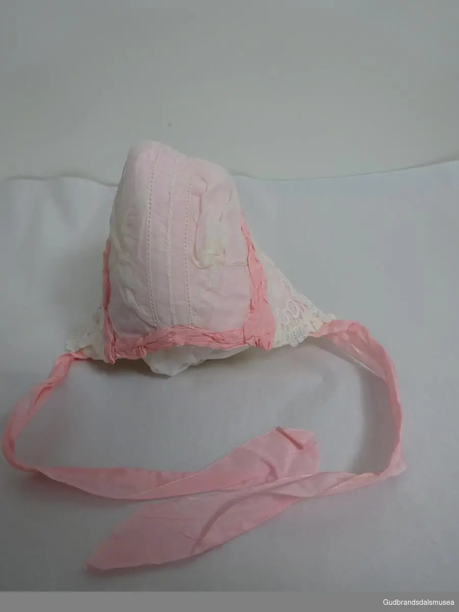 Dåpslue. Rosa med hvite blonder, rosa pynte- og knytebånd. 
Følger med en håndskrevet lapp med "Dåpsluve"