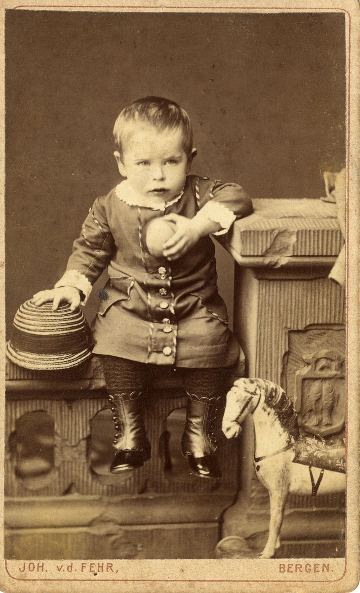 Portrett av et barn med ball og lekehest, sittende på kulissgjerde.