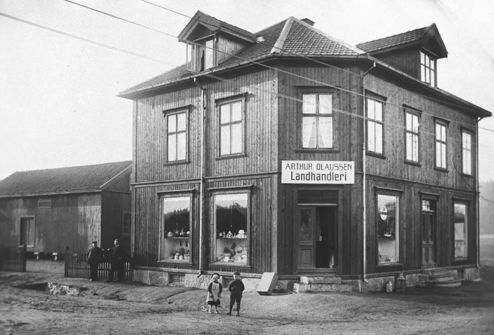 På dette foto av bygningen Aasheim fra 1915 ses Arthurs datter Ruth og sønnen Asbjørn foran landhandelen. Dette var før det ble manufakturbutikk i hjørnedelen av bygningen. På fotoet kan man se en lagerbygning ved siden av landhandelen. Foto: MiA.