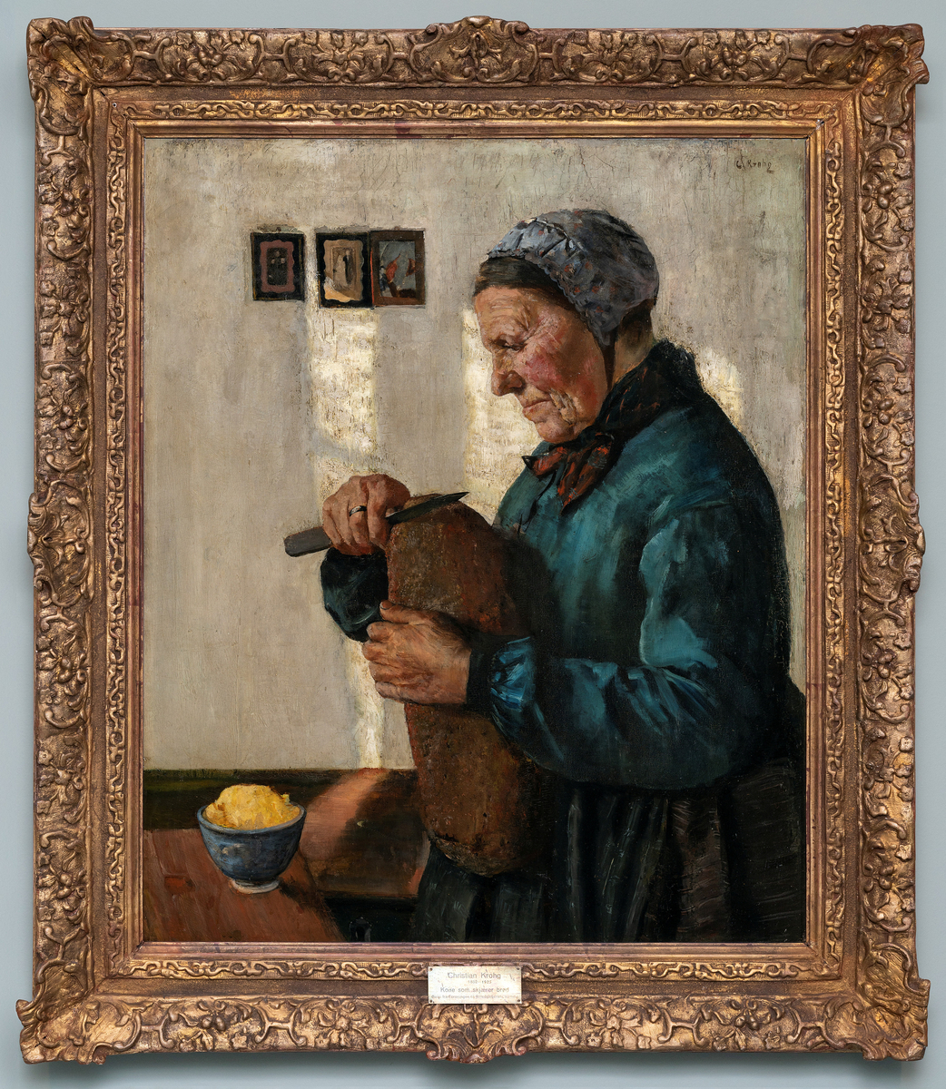 Eldre kvinne i blå bluse og brunt skjørt ved et bord i ferd med å skjære et brød som hun holder foran brystet. Blå kopp med smør på bordet.