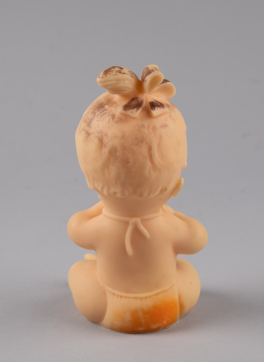 Leketøy av mykplast i form av en sittende baby. Den har orange bleie og smekke, blåe øyer og håret er satt opp i en sløyfe.