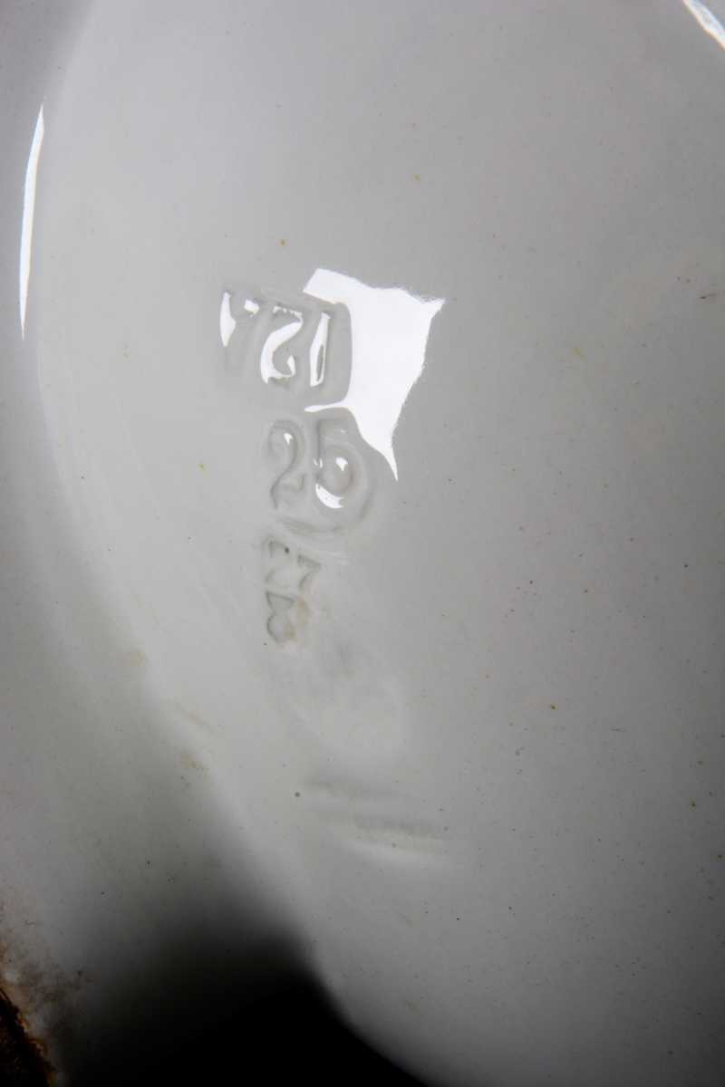 Hjørnevask av hvitt porselen. Den har rund skål og to rette sider med høy kant, slik at vasken passer i et hjørne. Vasken har sluk og rør av metall. På underiden av skålen er produksjonsstempel til firmaet "Johnson Brothers" med en ørn som holder et våpenskjold med initialene "JB", flankert av seks britiske flagg.