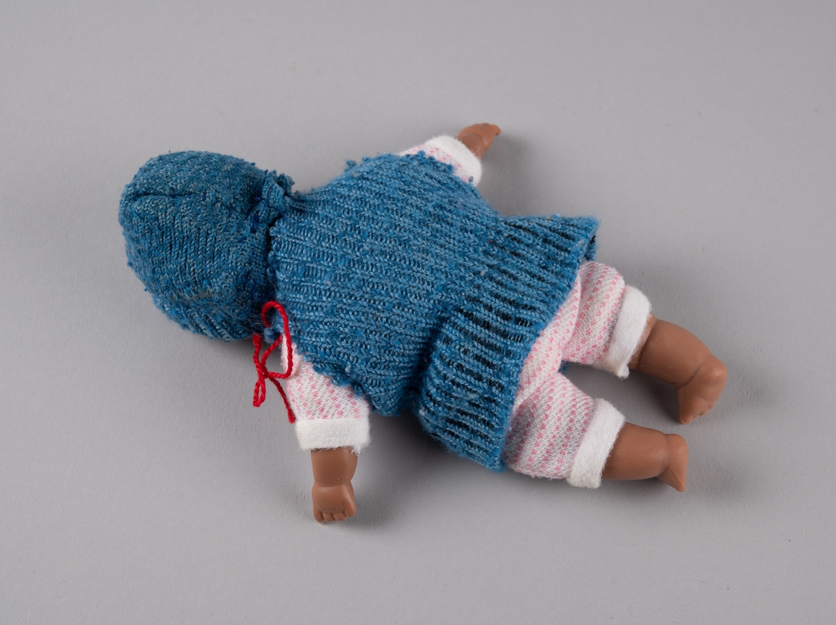 Babydukke med hode, armer og føtter av plast, og torso av tekstil fylt med vatt. Den er kledd i hjemmestrikket, rosa og hvit buksedrakt, samt blå genser og lue. Lua har en rød hakesnor.