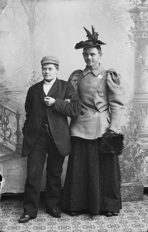A woman in men's clothes and a man in women's clothes. The man has a moustache, and holds the woman under his arm.