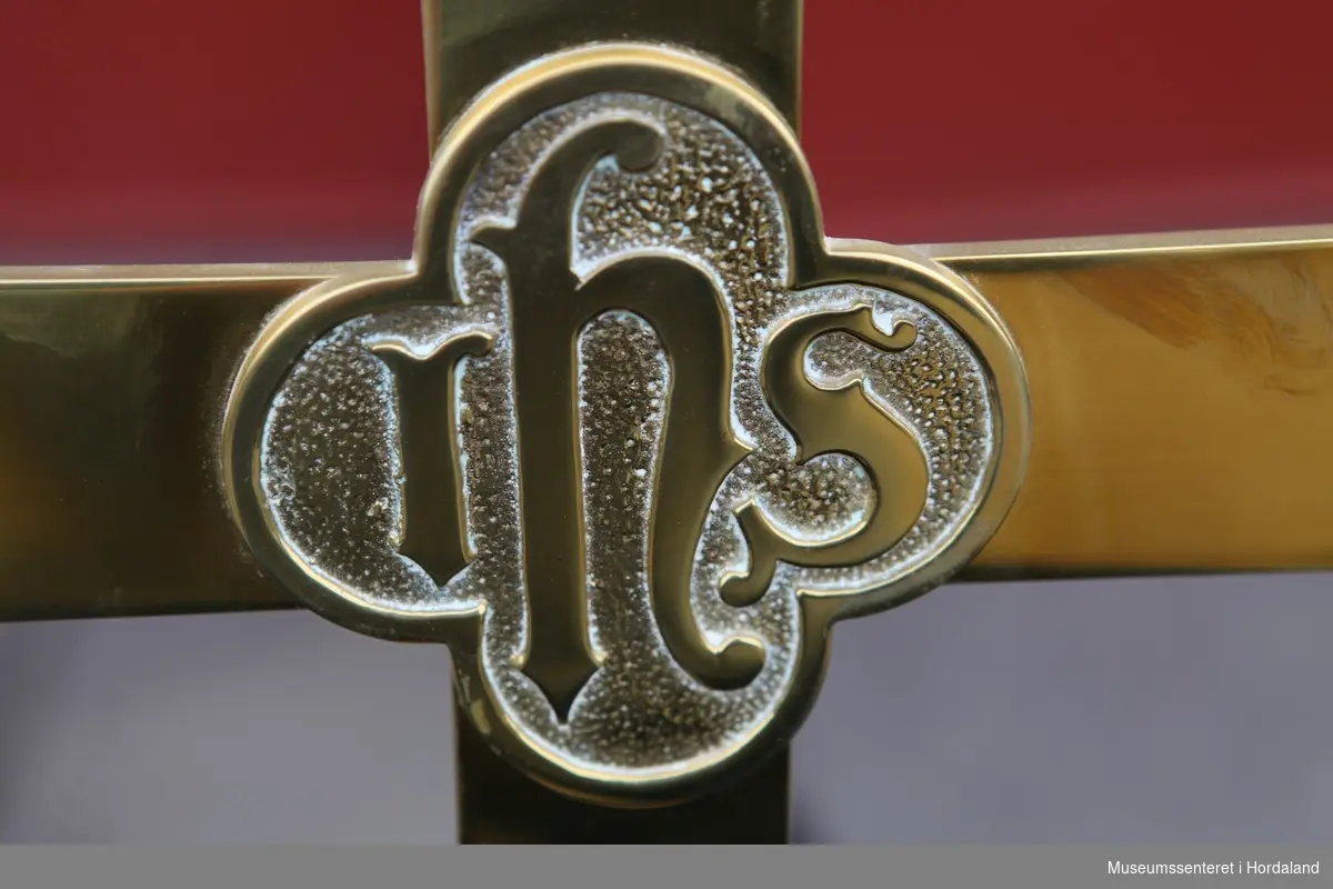 Alterkors i messing. Inngavert på midten av korset står "IHS" i en firearmet rosett. Korset står på en kvadratisk fot.