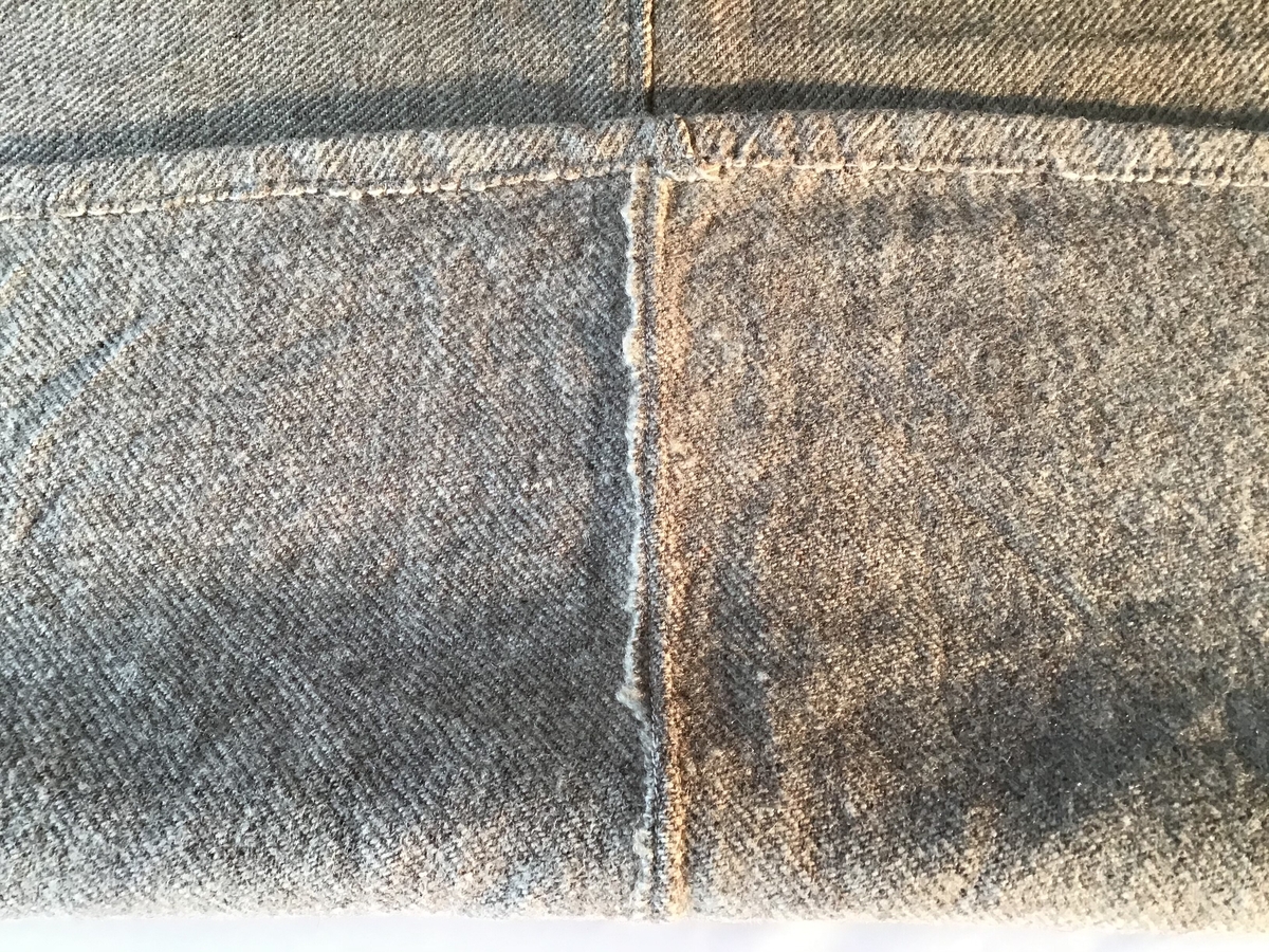 Krypertvevd ullteppe av gråmelent garn. Teppet er vevd i to deler som er sydd sammen på langs. Hver ende har en smal fold.