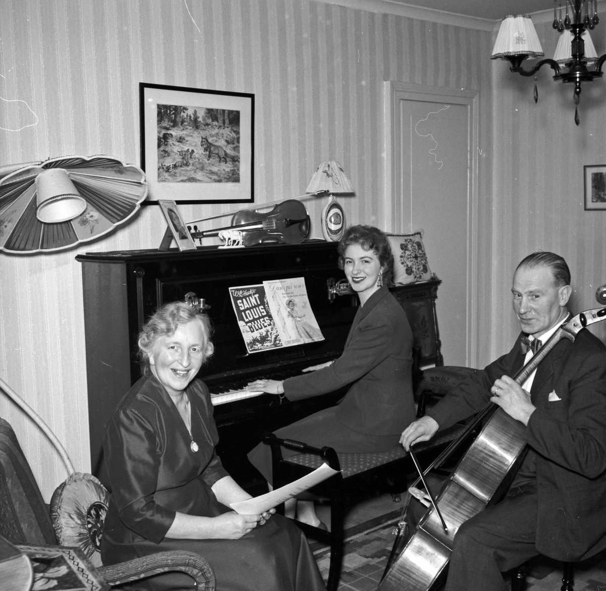 Äldre kvinna med sånghäfte, yngre kvinna vid piano samt äldre man med kontrabas (basfiol)