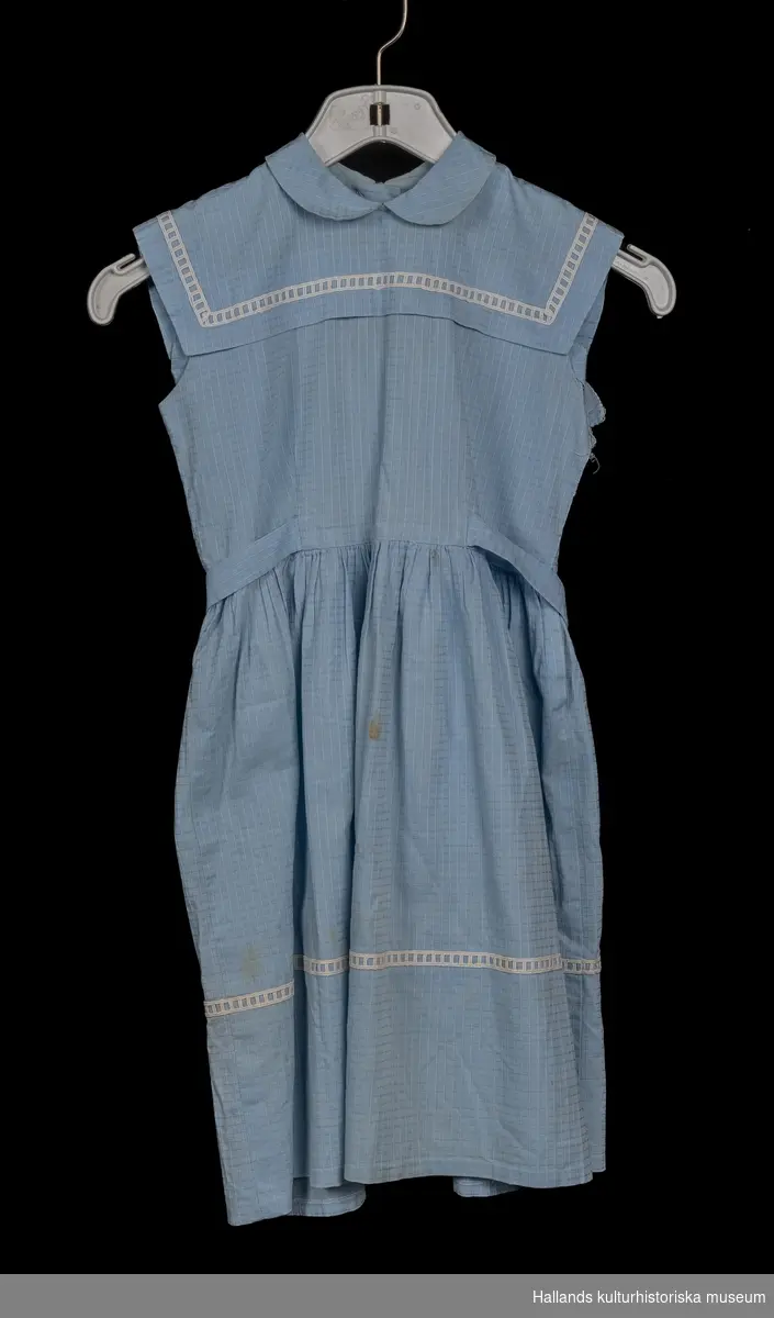 Blå bomullsklänning. Vit spets dekor på kragen och botten av kjolen. Tyget är i blå bomull med rutigt mönster.