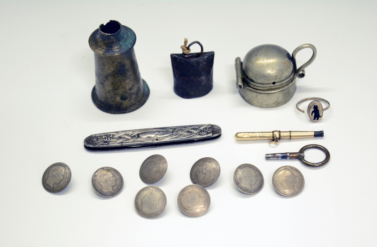 Dreid eske av tre med diverse metallgjenstander i: 7 knapper laget av mynter, ring, lokk, flaske, bjelle, lommekniv og to urnøkler.
