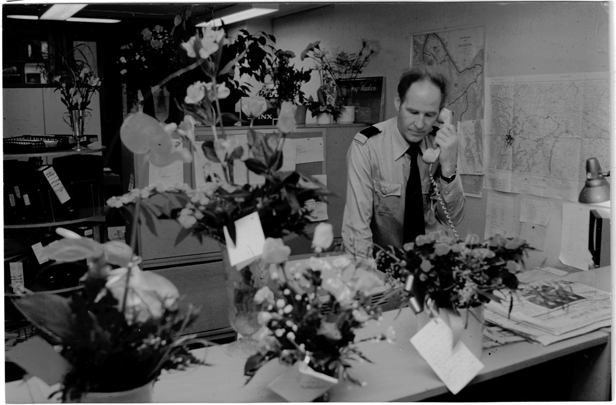 En uniformert politimann snakker i telefonen stående bak et skrivebord. Mannen er omgitt av mange blomsterbuketter.