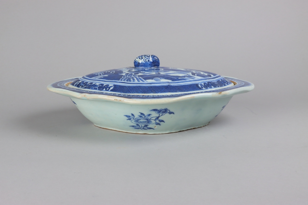 The lid with an oval slightly domed form, on top a knob shaped as a fruit. On the lid landscape scenes of pagodas, buildings,  bridges, figures with parasolls,  gardens and waters. The edge of the lid is decorated with a wide dark blue border with a criss cross pattern and reserves filled with symbols of good fortune.. The outside of the dish is decorated with rose branches. All decor in blue underglaze. The base of the dish without glazing