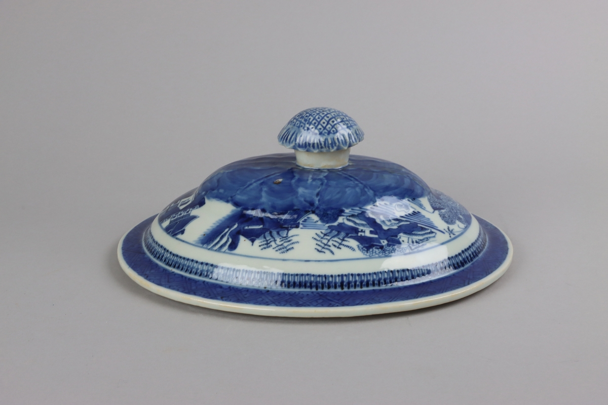 Oval shaped and slightly domed with knob in form of a flower head. On the top of lotus leaves and a list of pagoda lanscapes. The edge of the lid decorated with a dark blue  border in a criss cross pattern. All decor in blue underglaze. The inside of the lid is without decorations.