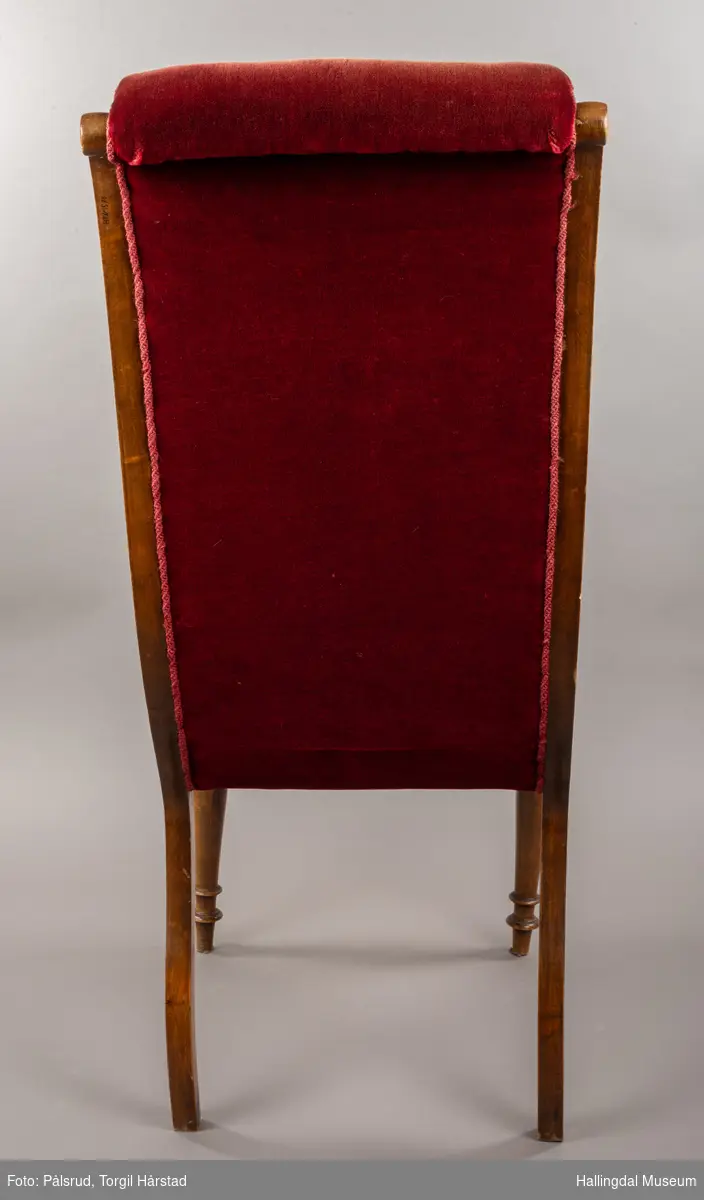 En kaminstol i rød fløyel. Langs setekantene er det montert lys røde tvinete bånd. Stolbeinene er i lyst tre. De bakerste beinene går oppover og støtter ryggen i en bue. De fremste beinene er dreid.