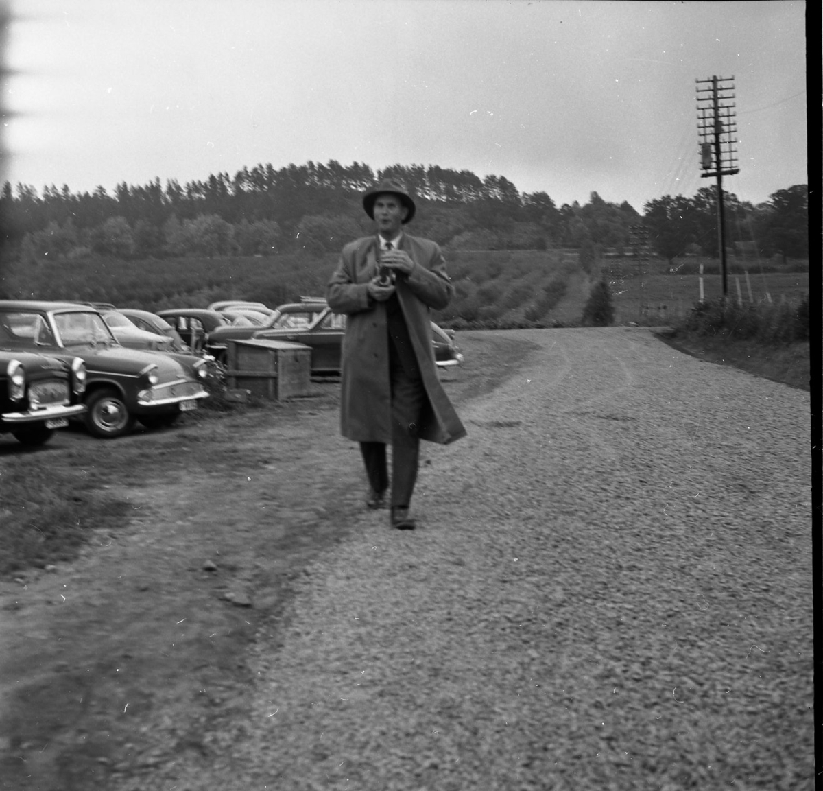 En man går längst en väg. Han håller i en kamera.  Bredvid vägen står bilar parkerade.