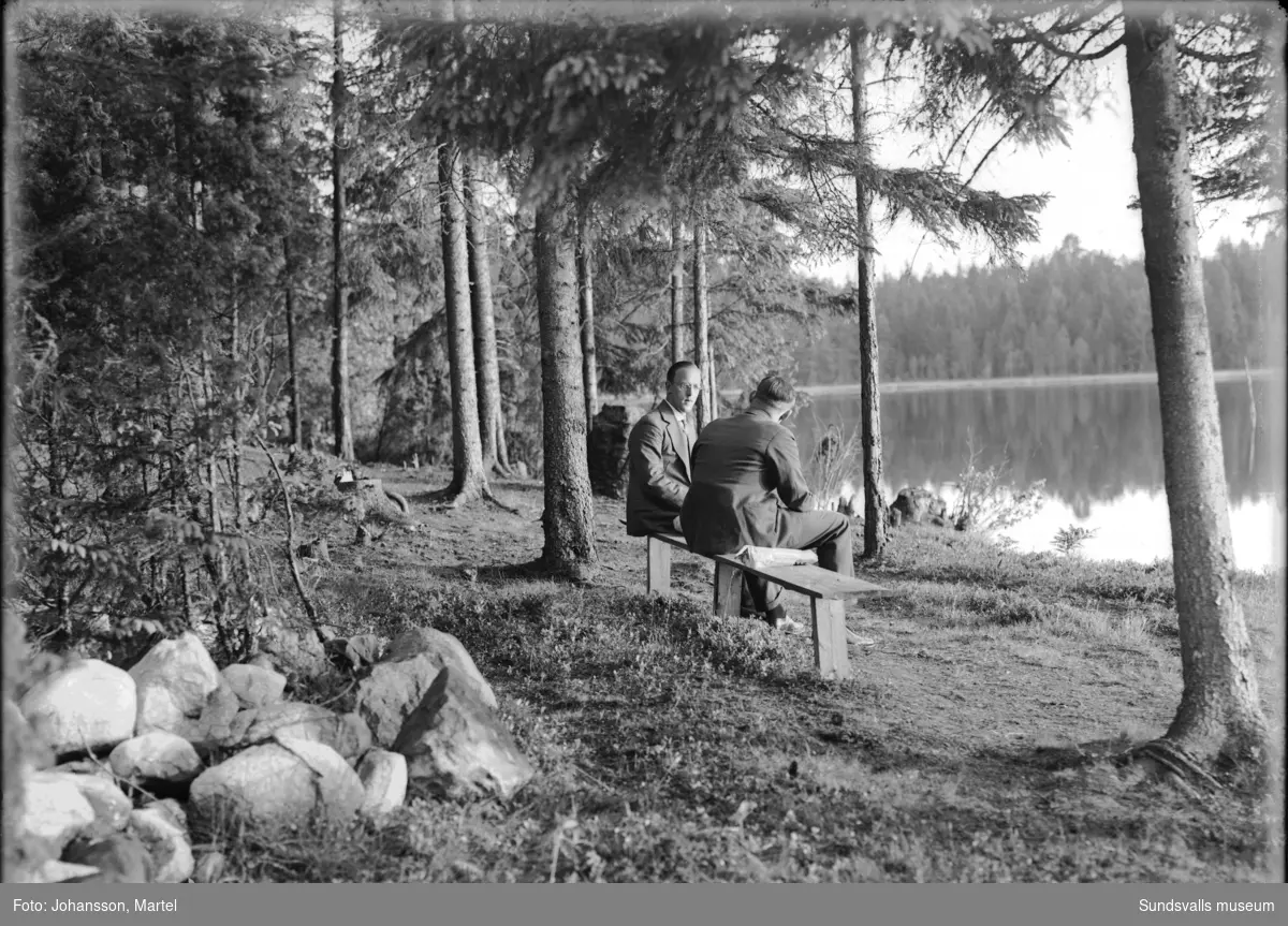 Gruppbild i det gröna med män, kvinnor och barn. Tvåa från vänster är fotografen själv, Martel Johansson. På den andra bilden sitter två av männen på en bänk invid en skogssjö.