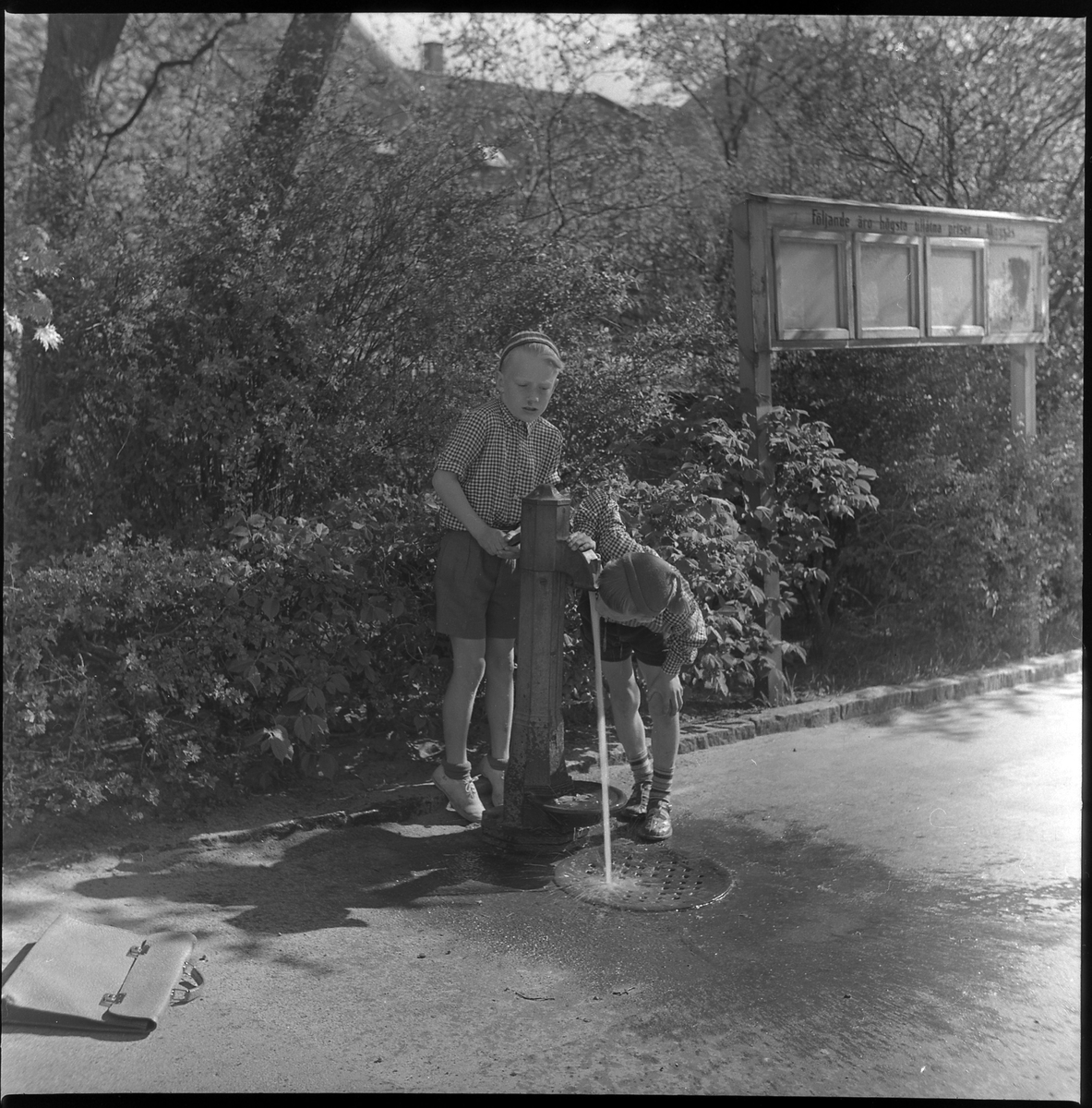 Två pojkar använder pumpen vid Lilla torget. Maj 1950. I bakgrunden ett skyltskåp med texten: "Följande äro högsta tillåtna priser i Alingsås".