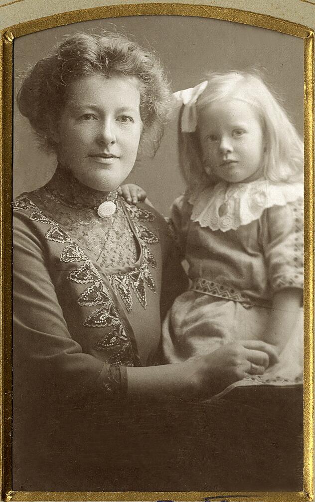En kvinna i höghalsad klänning med brosch i halsgropen, tillsammans med sin dotter i klänning med stor vit krage.
Midjebild, Ateljéfoto.
