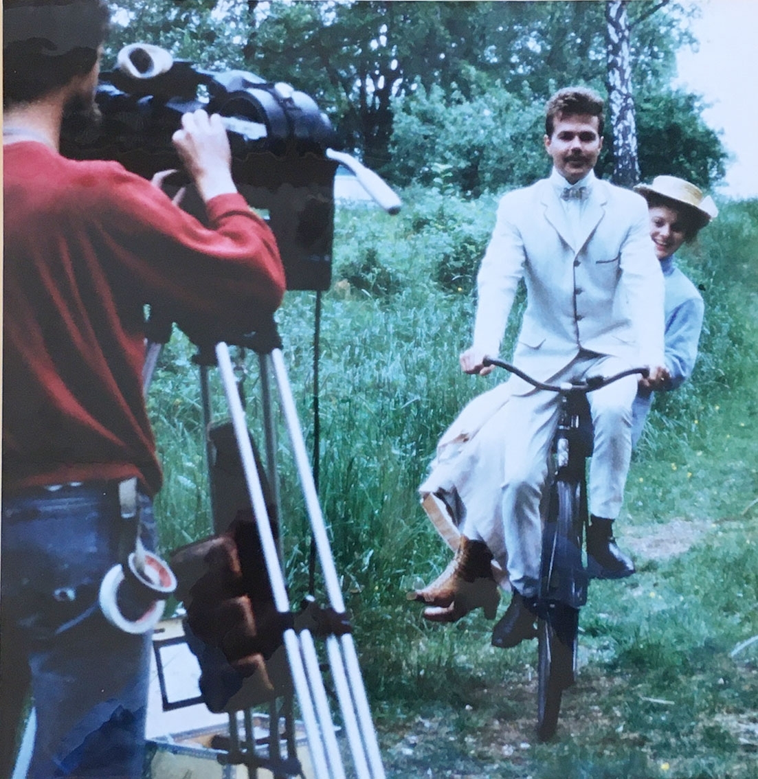 En man i ljus kostym på en cykel med en kvinna i hatt och ljus kappa på pakethållaren, de skrattar. De cyklar mot en man med filmkamera på stativ som filmar dem.