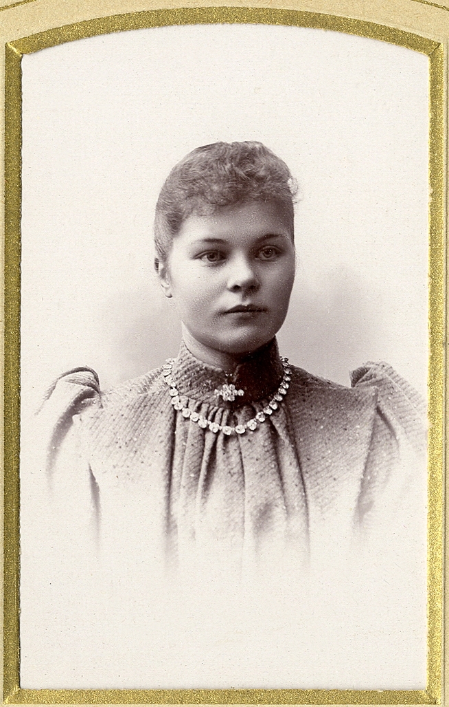 En okänd kvinna i höghalsad ljus klänning med en brosch i halsgropen och halsband.
Bröstbild, halvprofil. Ateljéfoto.