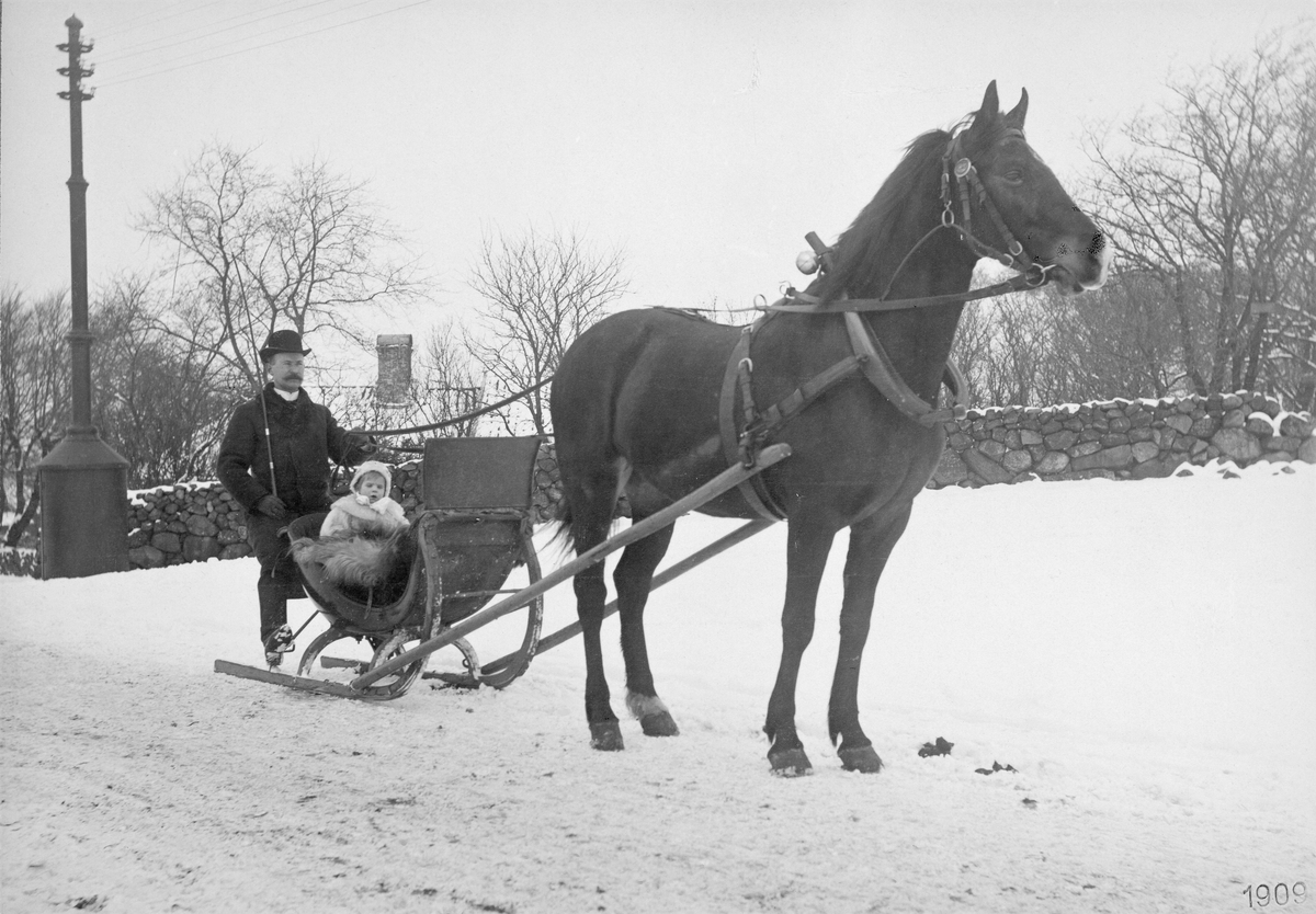 Hästekipage i snö med ett barn i släden och en kusk med piska. Bilden tagen utmed Kungsgatan, troligen är det södra kyrkogården som syns i bakgrunden. (Se även bild MR2_1027)