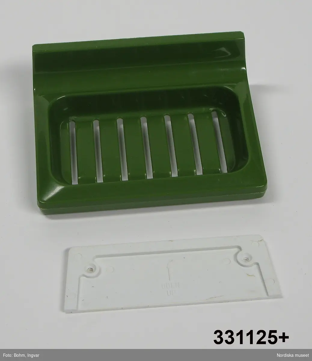 Tvålhållare, av grön (avocadogrön) formgjuten plast.  Löst vitt bakstycke/monteringsplatta med text "OBEN" och en pil. 
/Johan Åkerlund 2010