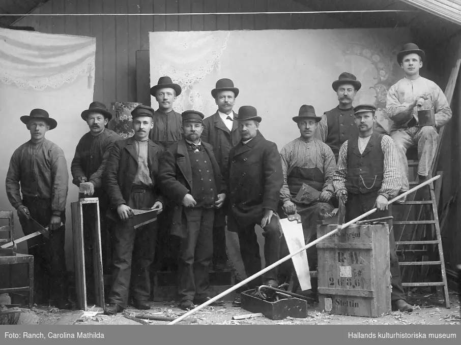 Ur Familjen Nilssons fotosamling. Gruppbild av snickare och målare, anställda hos byggmästare Johannes Nilsson som själv syns iklädd kubb som tredje person i nedre raden från höger.