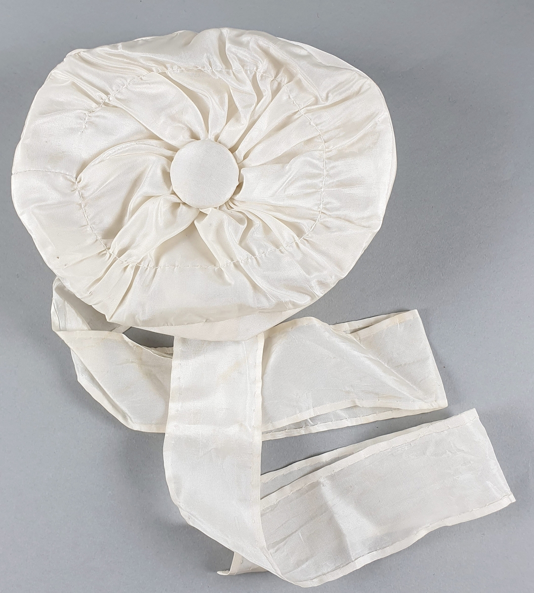 Hvit babylue av silke med lange bånd til å knyte under haken. Stor knapp på toppen og bånd rundt kanten.