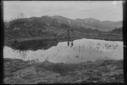 To menn stående ved lite vann i fjellandskap. Fjelltopper i 