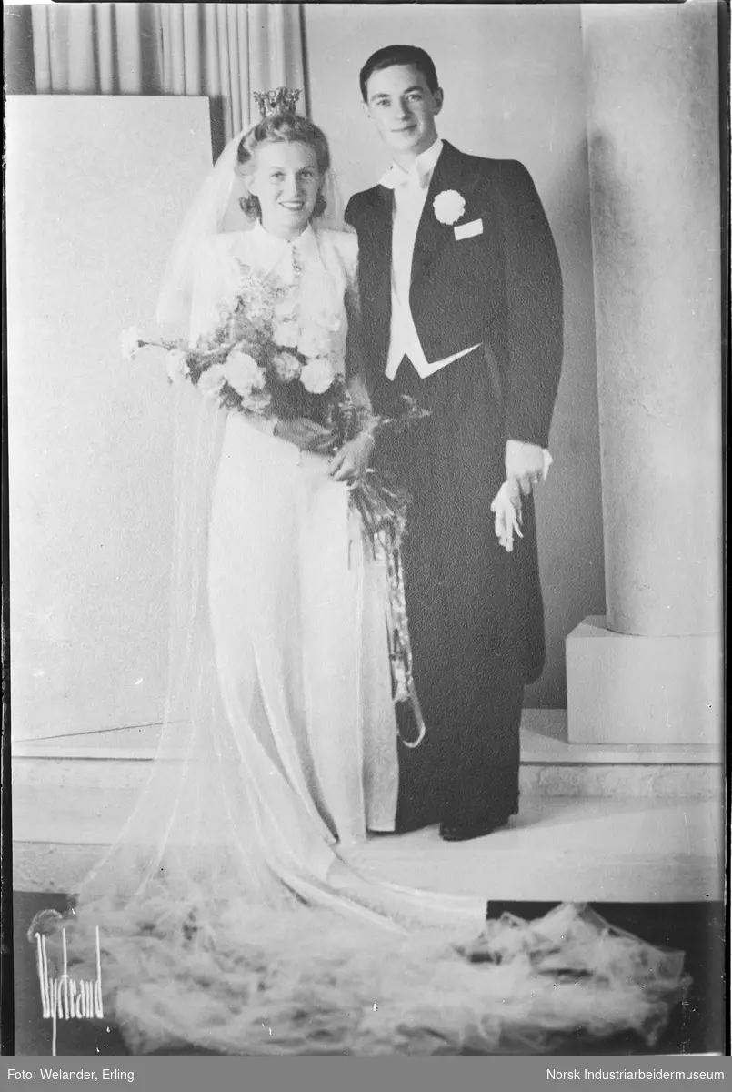 Reprofotografert brudebilde. Brudepar stående i studio. Brud i brudekjole, slør, krone på hodet og brudebukett i hendene. Brudgommen i kjole og hvitt med hansker i hånden.