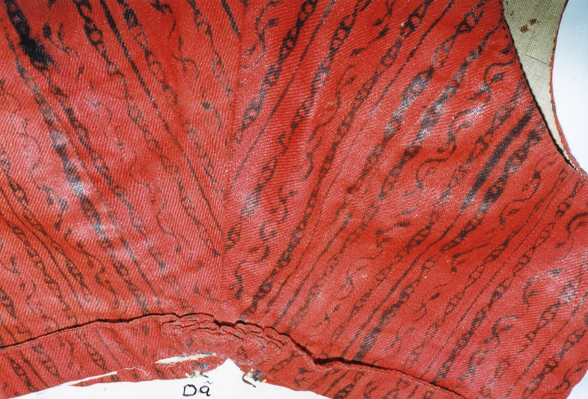 Livstycke, i kort modell med 3 cm bred uppvikt nederkant, av röd glättad yllekypert med tryckt mönster i svart linoljefärg i smala ränder och bladslingor. Två framstycken/sidostycken med djup urringning som går upp i en snibb mitt fram, utmed urringningen framtill och upp till axeln ett smalt svart sammetsband, smala axelband, knäpps med sju hyskor och hakar av järntråd (flera saknas). Två ryggstycken avsmalnande nedåt i tre uppåtvikta skörtveck. Foder av vit linnelärft. 
Har tillhört givarens mormor Maria Pehrsdotter 1796-1859 och har förmodligen använts tillsammans med bröllopsbandet DR 0583.
 
Flera livstycken och västar i rött ylle med svart oljefärgstryck i flera mönstervarianter finns från Kronobergs län, sannolikt tryckta av en lokal tygtryckare.
Jfr. Västar NM 0077706b, NM 0077707b, DR 0051,livstycke NM 0026764

U.4.21. Ingår i Textilinventeringen i Kronobergs län 1928-1930.

/Birgitta Blixt 2021.