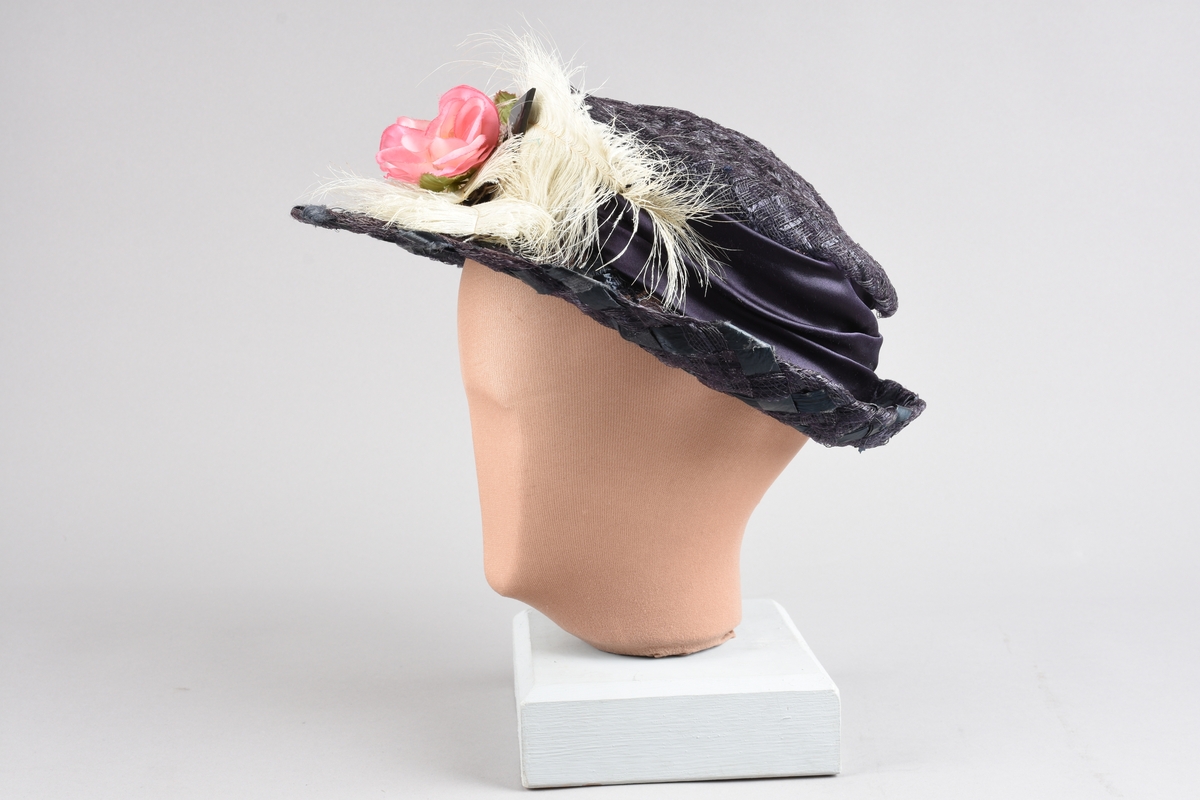 Mørkfiolett, flatpullet og bredbremmet hatt i  "bretonne-" el. "canotier-"fasong. Laget av nettvevede, kunstige stråbånd. Rundt pullen er et "hattebånd" av bred drapert sateng i samme farge, festet med spenne av hamret messingblikk. Senere laget om til kanutt-kostyme, med kunstig rosegren oppå offwhite strutsefjær dandert over pannen. Inni pullen er en skulderpute satt fast med sikkerhetsnål.  Hårnål og hatte-elastikk til feste av hatten.