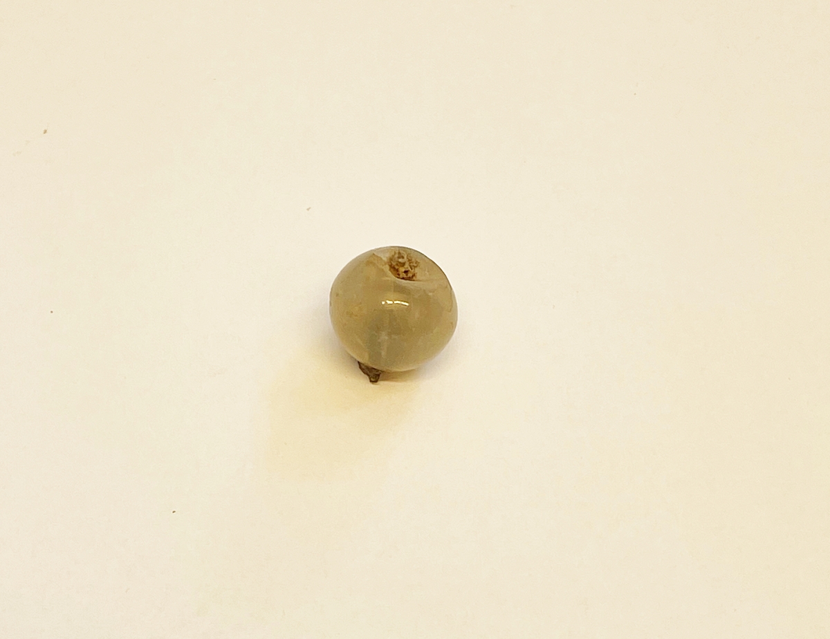 Rund knapp eller möjligen prydnadsnål av gråaktig, halvopak kalcedon. Öglan förrostad, men delvis bevarad inne i den genomborrade stenen. Har sannolikt prytts av metallornament på toppen.