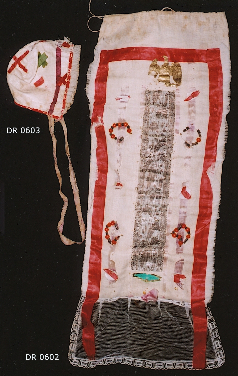 Rektangulär doppåse av småmönstrad  kattun i rosa och vitt, sprund i höger sidan upptill och dragsko med dragband av tvinnat lingarn, nertill kappa av spetskantad vit tyll.  Upptill och längs sidorna prytt med ett rött sidenband som hänger ner över kappan, innanför det ett smalt lila sidenband med påsydda små kransar av röda och svarta glaspärlor och röd/vita sidenbandskors, i mitten ett brett silverfärgat band med invävt flotterande blommönster krönt med en örn med utslagna vingar i guldpapper. Vid långsidorna en veckad tyllvolang. Foder av smalrandig bomullslärft i rosa och vitt.   
Jfr. DR 0604 Dä153 mössa i samma kattun.
 
Birgitta Blixt 2021.
