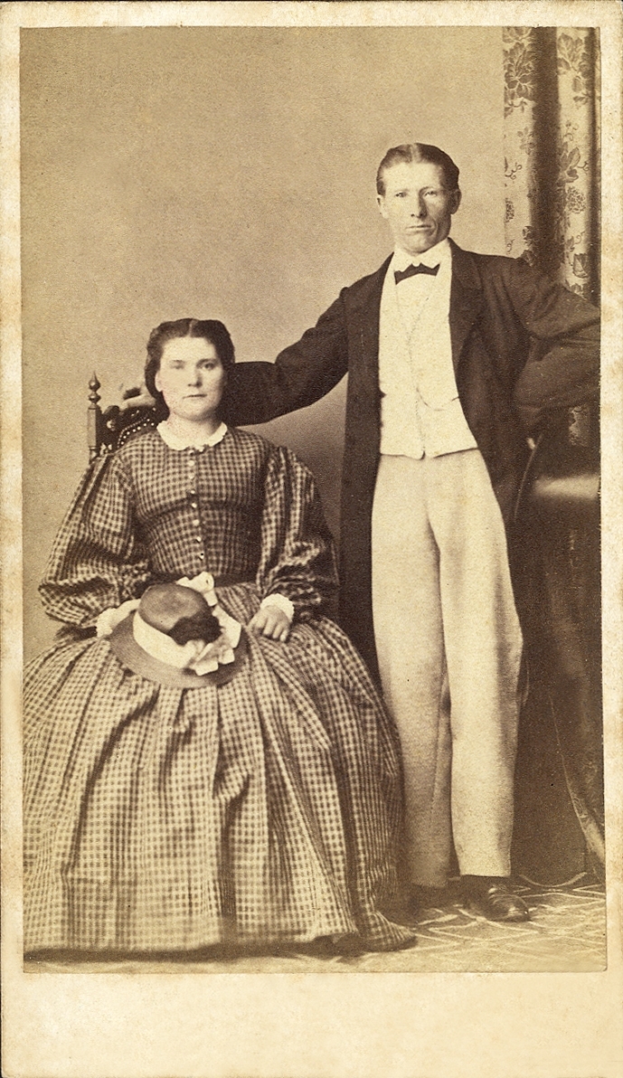 En kvinna i rutig krinolin tillsammans med en man i redingot och ljusa byxor.
På baksidan av fotot antecknat:"Jacobsson".
Helfigur. Ateljéfoto.