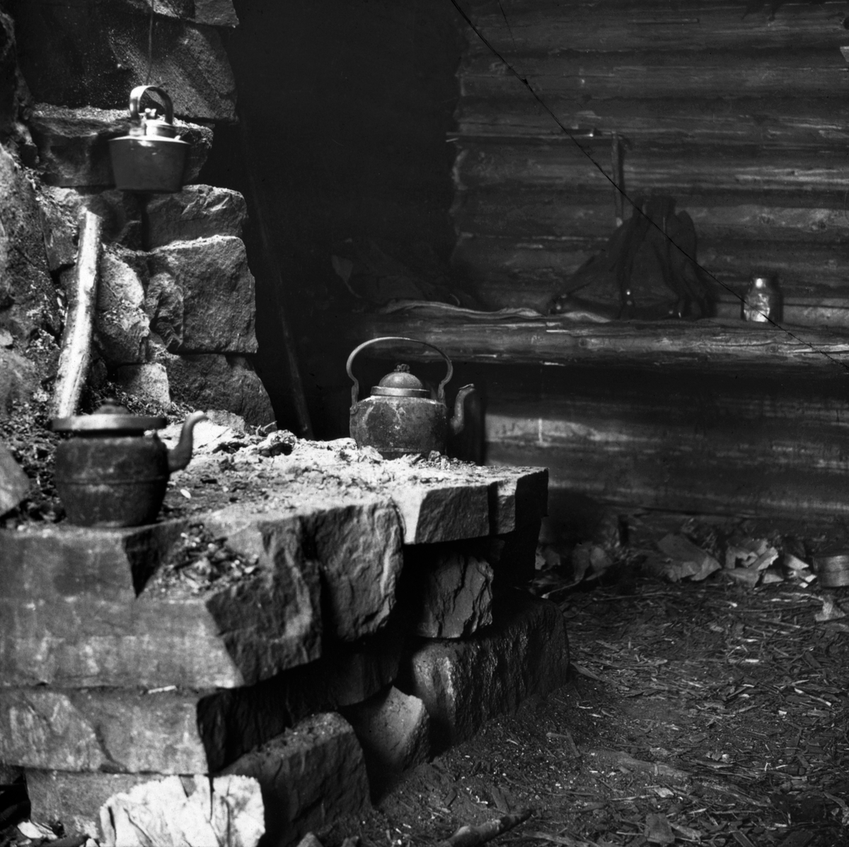 Interiør fra såkalt «murkoie» eller «peiskoie». Fotografiet er antakelig tatt i Hedmark eller Oppland i andre halvdel av 1920-åra. I forgrunnen til venstre ser vi det innredningselementet som var opphavet til de nevnte koietypebetegnelsene - en peis. Skogskoiene lå gjerne uvegsomt til i skogslandskapet, og de ble følgelig i hovedsak bygd av materialer man kunne finne på stedet, eller i hvert fall i nærheten av koietomta. Dette bildet er tatt i ei lita laftet stue, antakelig oppført av tømmer fra tørrgran eller annet virke som hadde liten kommersiell verdi. Inne i rommet var det sitte- og liggebrisker langs veggene, også her med rundtømmer som bærende elementer, men sannsynligvis med halvkløvninger eller bord i den flata skogsarbeiderne skulle sitte og sove på. Her sto det bare en bæremeis og ei lita blikkflaske på brisken. Peisen var oppført av kantete naturstein, antakelig med et minimum av bindemiddel. Den later til å være murt inntil en av tømmerveggene, og murverket skjermet i hvert fall veggen for ildsfarlige gnister. Den hadde dessuten et pipeløp som, i hvert fall når været var noenlunde vindstille, ledet mesteparten av røyken ut av rommet. Men mye av varmeenergien fra den brennende veden gikk også rett opp gjennom pipa - varmestrålinga inn i rommet var beskjeden. I det koierommet der dette fotografiet er tatt målte Sønnik Andersen likevel 32 grader 1,5 meter over jordgolvet tidlig på kvelden, mens karene lagde seg dagens varme måltid på peisen - etter tur. Samtidig var det bare 2 varmegrader 10 centimeter over golvet. Påfølgende morgen hadde romtemperaturen sunket til 14 grader, og nede ved golvet var temperaturen på frysepunktet. Dette forklares slik: «Her er peis. Den lave temperatur ved gulvet kommer meget av det. For at peisen skal trekke er det gjort hull under nederste kvarv i veggen. Døren er gissen omkring karmene og her er jordgulv. Alder 10 år.»

I peisen vi ser på dette bildet var det ikke mindre enn tre kaffekjeler. To av dem stå ytterst på den såla der ilden skulle tennes, den tredje hang i en ståltråd høyere oppe, der varmestrålinga var på det kraftigste. Johan Sønnik Andersens far, fylkeslege Einar Andersen (1864-1954), skrev en epilog til sønnen Sønnik Andersens «Undersøkelse av skogshytter og skogsarbeidernes levevilkår». Fylkeslegens tekstsekvens handlet om skogsarbeidernes kosthold. Når det gjaldt kaffe fomulerte distriktslegen seg balnt annet slik: «Om vinteren er det ikke lyst nok til å arbeide i skogen lenger enn fra 7 ½ fm. til kl. 5 em. De må utnytte arbeidstiden og får ikke tid til å få sig ordentlig kokemat før dagsarbeidet er endt. Hele arbeidsdagen lever de på kaffe og smørbrød. Når de så kommer inn kl. 5 om eftermiddagen, må de foreløpig stagge den verste sult – fremdeles med kaffe og smørbrød.» Men etter hvert lagde de seg vanligvis et varmt måltid, etter tur og etter fattig evne. Andersen føyer til denne betraktningen om kaffen: «Der drikkes svært meget kaffe i kojene – 5 ganger om dagen foruten en del ganger om natten, og visstnok store porsjoner hver gang. Det er forståelig at folk som arbeider i kulden, har trang til varmt drikke og noget å stive sig op på. Men all denne kaffen kan allikevel ikke være bra.»

Fotografiet er tatt i forbindelse med undersøkelser forstkandidat Johan Sønnik Andersen gjorde i Hedmark, Oppland og Nord-Trøndelag i andre halvdel av 1920-åra.  Observasjonene og refleksjonene hans ble utgitt under tittelen «Undersøkelse av skogshytter og skogsarbeidernes levevilkår» i 1932.  Norsk Skogmuseum har overtatt en del av de fotografiske negativene fra Sønnik Andersens undersøkelser i Hedmark, Oppland og Trøndelag.  Noen få av disse er brukt som illustrasjoner i det nevnte heftet.  De er imidlertid – antakelig for å sikre involverte skogeiere og skogsarbeidere en viss anonymitet – svært knapt tekstet fra opphavsmannens side.  Dette har naturligvis også fått konsekvenser for hvordan Norsk Skogmuseum kan registrere motivene i sin fotodatabase.